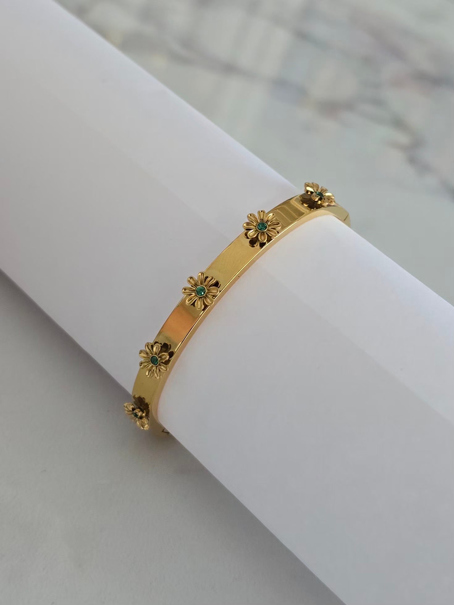 Bracelet jonc Margarita doré avec cinq fleurs 3D aux détails verts sur présentoir blanc.