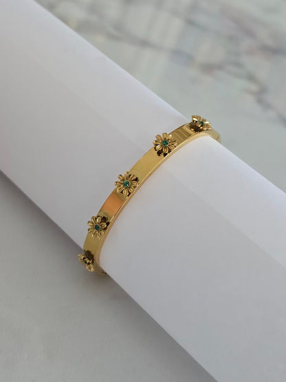 Bracelet jonc Margarita doré avec cinq fleurs 3D aux détails verts sur présentoir blanc.