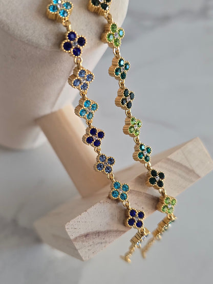 Bracelets Princesa dorés suspendus sur présentoir, ornés de délicates fleurs à strass bleus et verts