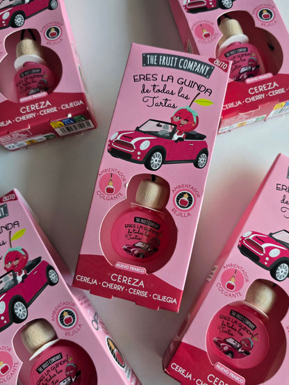 Parfum voiture senteur cerise The Fruit Company présenté dan son emballage rose avec plusieurs diffuseurs en arrière-plan.